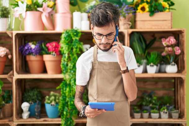 Молодий іспаномовний чоловік флорист розмовляє по смартфону, використовуючи тачпад у квітковому магазині
 - Фото, зображення