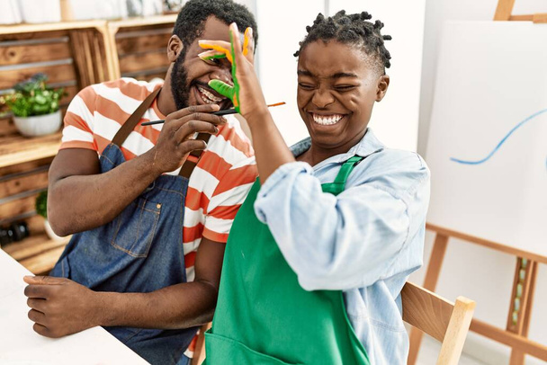 Αφροαμερικανοί ζωγράφοι ζευγάρι χαμογελώντας χαρούμενα χέρια ζωγραφική κάθεται στο τραπέζι στο στούντιο τέχνης. - Φωτογραφία, εικόνα
