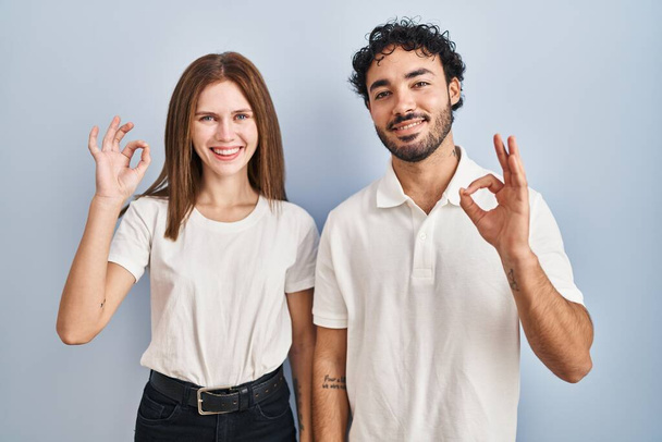 Mladý pár nosí ležérní oblečení stojí spolu s úsměvem pozitivní dělat v pořádku znamení s rukou a prsty. úspěšný výraz.  - Fotografie, Obrázek