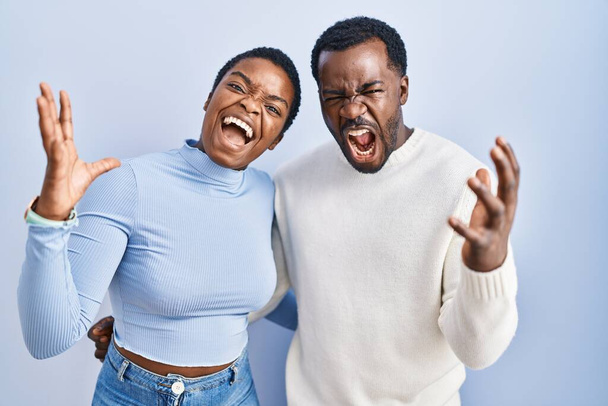 Young african american couple standing over blue background crazy and mad shouting and yelling with aggressive expression and arms raised. frustration concept.  - Fotografie, Obrázek