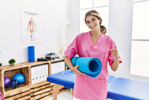 Young physiotherapist woman working at pain recovery clinic holding yoga mat smiling happy pointing with hand and finger  - Valokuva, kuva
