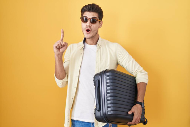 Yaz tatiline giden bavulu tutan genç İspanyol adam başarılı bir fikirle işaret ediyor. Heyecanlı ve mutlu. Bir numara..  - Fotoğraf, Görsel