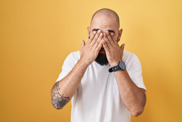 Young hispanic man with beard and tattoos standing over yellow background rubbing eyes for fatigue and headache, sleepy and tired expression. vision problem  - Foto, Imagem