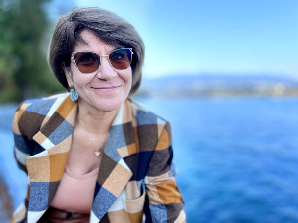 dospělá žena v slunečních brýlích v kostkovaném kabátě sedí na břehu Tichého oceánu v západoindickém parku má krátké vlasy husté Usměje se a směje se do rámu očima - Fotografie, Obrázek