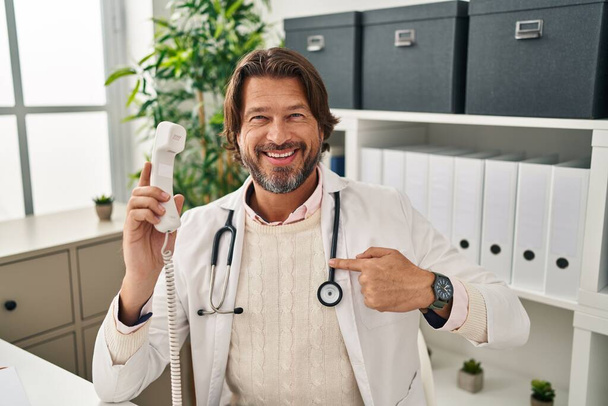 Bell'uomo medico di mezza età che parla al telefono alla clinica indicando un sé sorridente felice e orgoglioso  - Foto, immagini