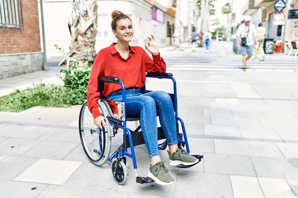 Young brunette woman sitting on wheelchair outdoors doing ok sign with fingers, smiling friendly gesturing excellent symbol  - Фото, зображення