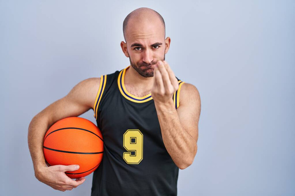 Молодой лысый мужчина с бородой в баскетбольной форме держит мяч делая итальянский жест с рукой и пальцами уверенное выражение  - Фото, изображение