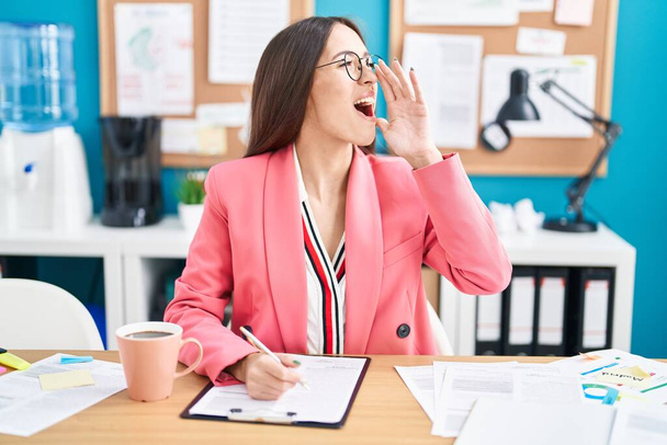 Giovane donna ispanica che lavora in ufficio indossando occhiali urlando e urlando forte a lato con la mano sulla bocca. concetto di comunicazione.  - Foto, immagini