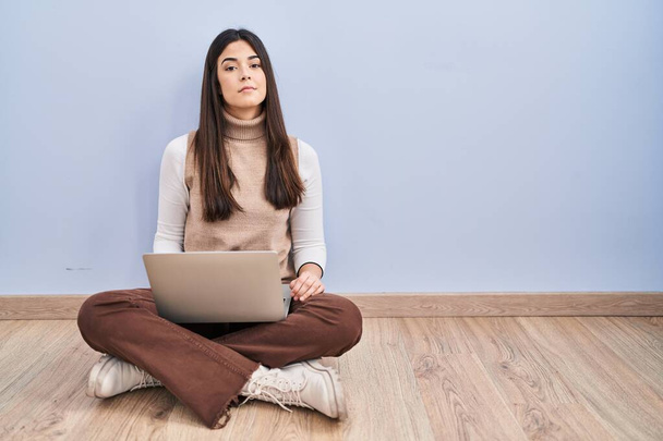 Young brunette woman working using computer laptop sitting on the floor relaxed with serious expression on face. simple and natural looking at the camera.  - Zdjęcie, obraz