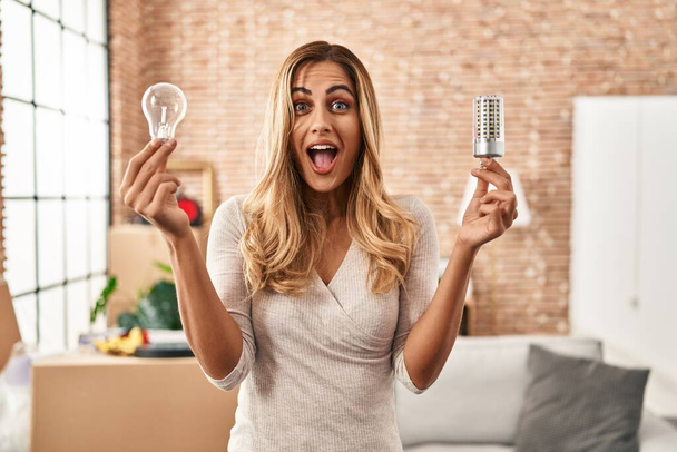 Young blonde woman holding led lightbulb and incandescent bulb celebrating crazy and amazed for success with open eyes screaming excited.  - Fotó, kép