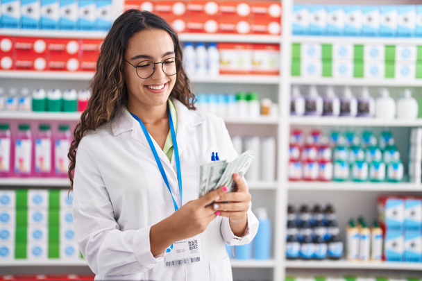 Giovane bella donna ispanica farmacista sorridente fiducioso contando dollari in farmacia - Foto, immagini