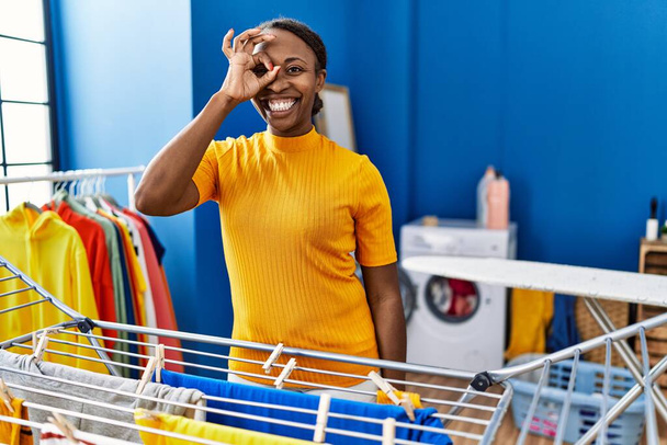Mujer africana colgando ropa en el tendedero sonriendo feliz haciendo bien signo con la mano en el ojo mirando a través de los dedos  - Foto, Imagen