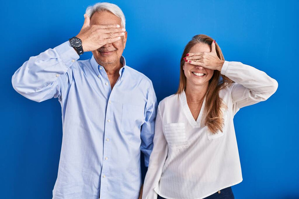 青い背景の上に立っている中年のパニックカップルは笑顔で、驚きのために目を覆う顔を手で笑っています。視覚障害の概念.  - 写真・画像