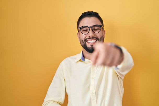 Hispánský mladý muž v obleku a brýlích, smějící se vám, ukazující prstem na kameru s rukou přes tělo, výraz studu  - Fotografie, Obrázek