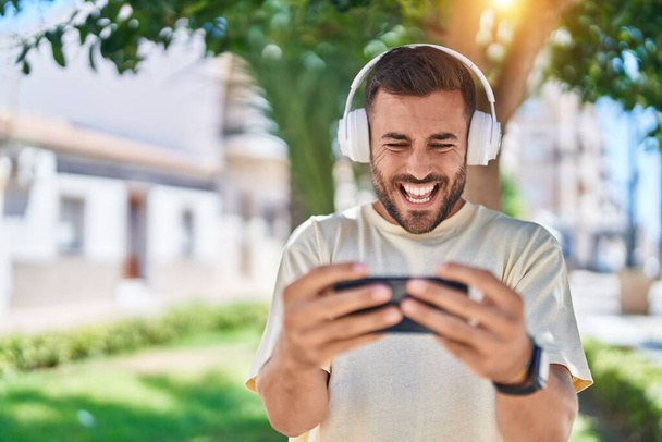 Joven hombre hispano sonriendo confiado jugando videojuego en el parque - Foto, imagen