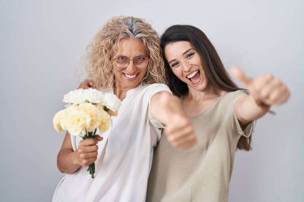 Мати і дочка тримають букет білих квітів, схвалюючи позитивний жест рукою, великі пальці посміхаються і щасливі за успіх. жест переможця
.  - Фото, зображення