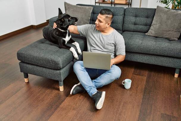 Young hispanic man using laptop sitting on the floor with dog at home. - Фото, зображення