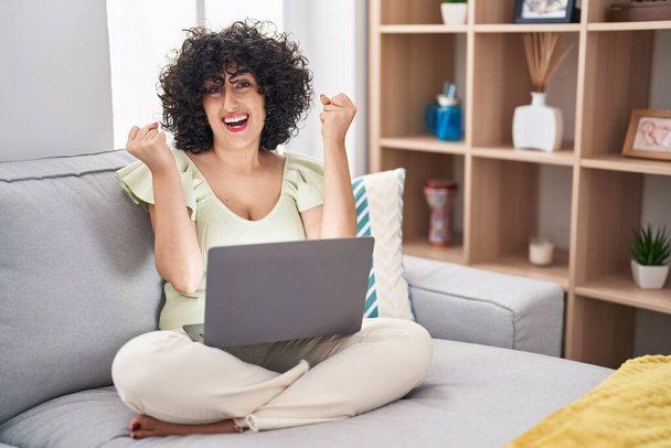 Jeune femme brune avec les cheveux bouclés à l'aide d'un ordinateur portable assis sur le canapé à la maison célébrant surpris et étonnés pour le succès avec les bras levés et les yeux ouverts. concept gagnant.  - Photo, image