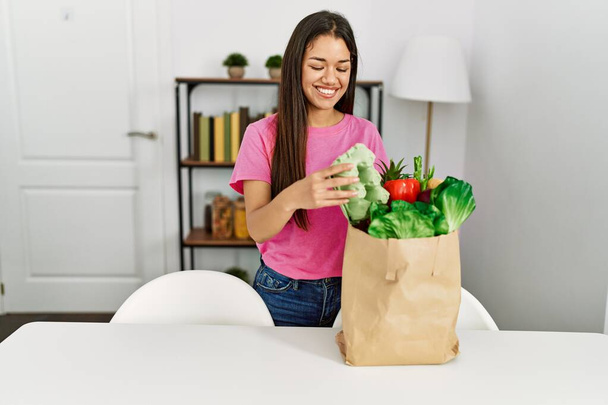 Молодая латиноамериканка держит дома еду из пакета с продуктами - Фото, изображение
