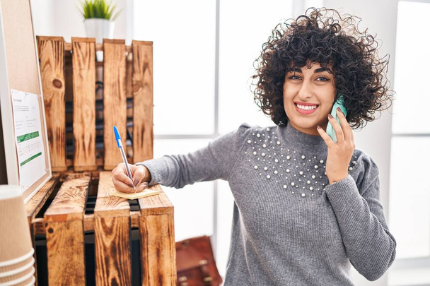 Junge Geschäftsfrau aus dem Mittleren Osten spricht im Büro auf dem Smartphone und schreibt auf ein Erinnerungsblatt - Foto, Bild