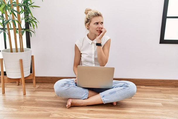 Mulher loira jovem usando computador portátil sentado no chão na sala de estar olhando estressado e nervoso com as mãos na boca mordendo unhas. problema de ansiedade.  - Foto, Imagem