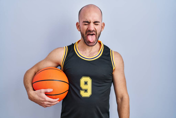 Mladý plešatý muž s plnovousem v basketbalové uniformě, držící míček, vystrkující jazyk, šťastný s legračním výrazem. koncepce emocí.  - Fotografie, Obrázek