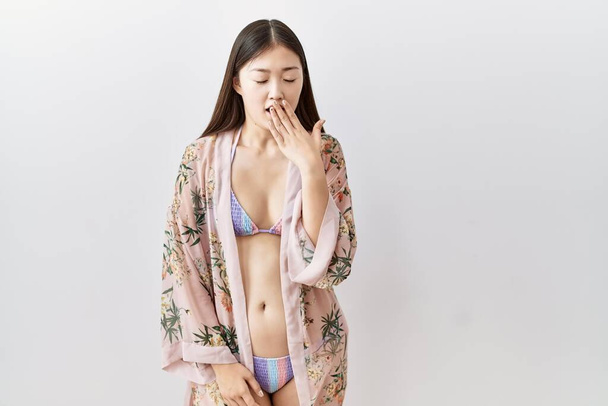 Молодая азиатка в бикини и цветочном кимоно скучает зевая усталая закрывая рот рукой. беспокойство и сонливость.  - Фото, изображение