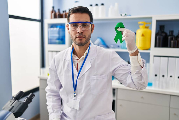 Joven hombre hispano con barba trabajando en laboratorio científico sosteniendo actitud de pensamiento de cinta verde y expresión sobria buscando confianza en sí mismo  - Foto, imagen