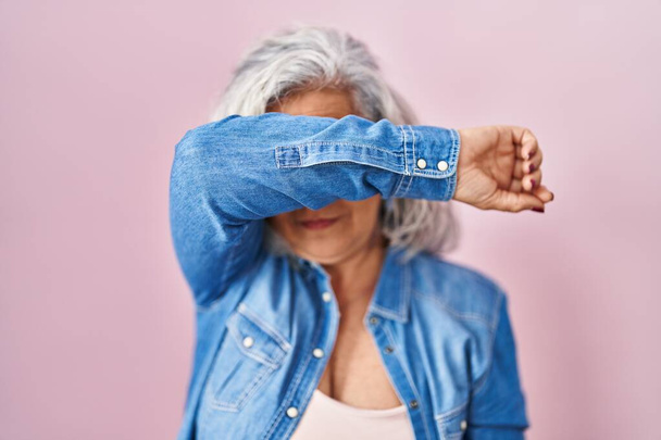 Middle age woman with grey hair standing over pink background covering eyes with arm, looking serious and sad. sightless, hiding and rejection concept  - Фото, изображение