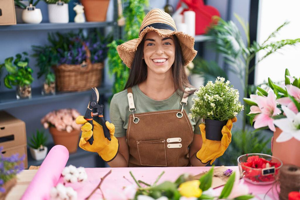 Die junge brünette Frau, die im Blumenladen arbeitet, lächelt mit einem fröhlichen und kühlen Lächeln im Gesicht. Zähne zeigen.  - Foto, Bild