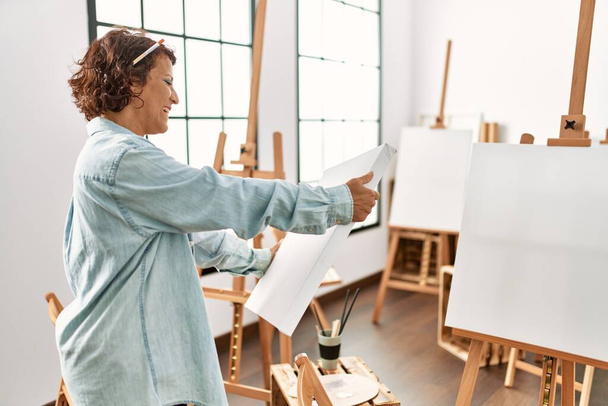 Middelbare leeftijd Latijns-Amerikaanse kunstenaar vrouw glimlachend gelukkig houden canvas in kunststudio. - Foto, afbeelding