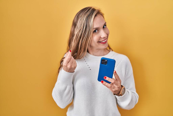 Młoda blondynka za pomocą smartfona wpisując wiadomość robi gest pieniądze z rąk, prosząc o wypłatę pensji, milioner biznesu  - Zdjęcie, obraz