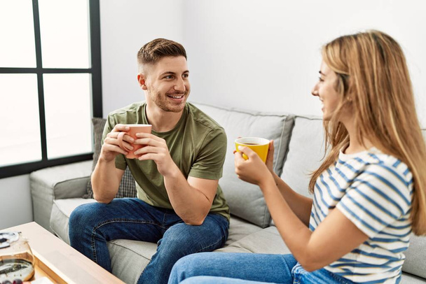 Jovem casal hispânico sorrindo feliz tomando café da manhã sentado no sofá em casa. - Foto, Imagem