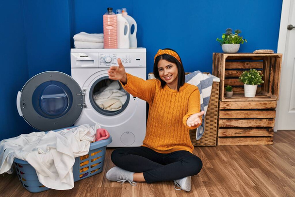 Junge hispanische Frau, die Wäsche wäscht und in die Kamera lächelt, mit offenen Armen zur Umarmung. fröhlicher Ausdruck, der das Glück umarmt.  - Foto, Bild