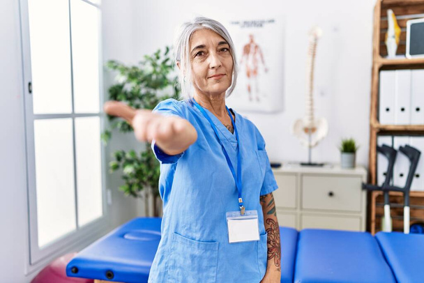 Седовласая женщина средних лет в форме физиотерапевта в медицинской клинике, улыбаясь дружелюбно предлагая рукопожатие в качестве приветствия и приветствия. успешный бизнес.  - Фото, изображение