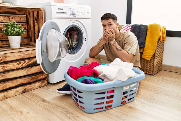 Молодой привлекательный мужчина кладет грязное белье в стиральную машину, шокируя закрывая рот руками за ошибку. секретное понятие.  - Фото, изображение