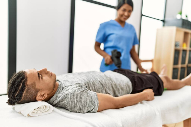 Junge lateinamerikanische Physioterapistin macht Reha-Behandlung für hispanischen Mann mit Massagepistole Perkussionswerkzeug in der Klinik. - Foto, Bild
