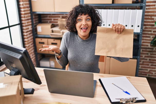 Latynoska lekarz kobieta z kręconymi włosami pracuje w małym biznesie e-commerce trzymając papierową torbę świętującą osiągnięcie ze szczęśliwym uśmiechem i wyrazem zwycięzcy z podniesioną ręką  - Zdjęcie, obraz
