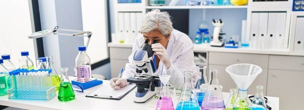Laboratuvarda mikroskop kullanan orta yaşlı kadın bilim adamı üniforması giyiyor. - Fotoğraf, Görsel