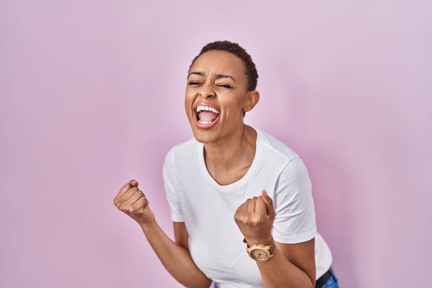 Schöne afrikanisch-amerikanische Frau steht über rosa Hintergrund sehr glücklich und aufgeregt und macht Siegergeste mit erhobenen Armen, lächelt und schreit nach Erfolg. Festkonzept.  - Foto, Bild