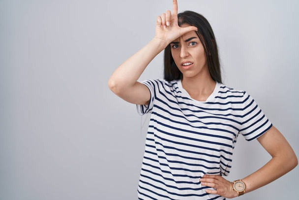 jonge brunette vrouw dragen gestreept t shirt maken plezier van mensen met vingers op het voorhoofd doen verliezer gebaar bespotten en beledigend.  - Foto, afbeelding