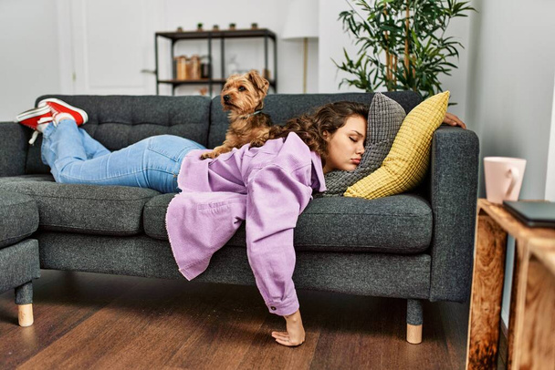Νεαρή όμορφη ισπανόφωνη γυναίκα ξαπλωμένη στον καναπέ και κοιμάται με σκύλο στο σπίτι - Φωτογραφία, εικόνα