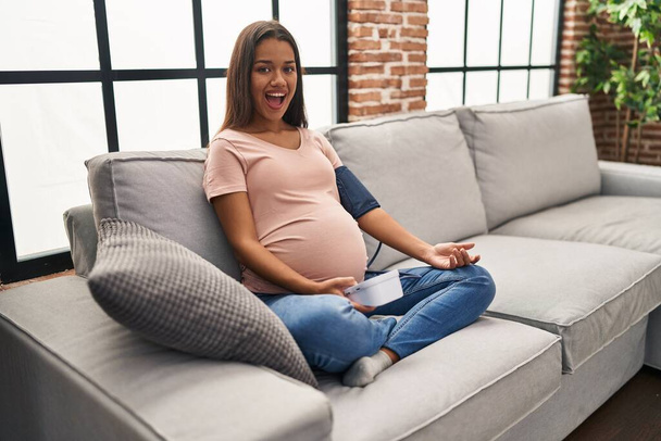 Jovem grávida usando monitor de pressão arterial sentado no sofá comemorando louco e surpreso com o sucesso com os olhos abertos gritando animado.  - Foto, Imagem