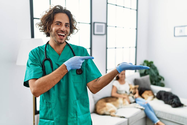 Mladý veterinární lékař muž kontrolu psů doma ohromen a usmívá se do kamery při prezentaci s rukou a ukazuje prstem.  - Fotografie, Obrázek