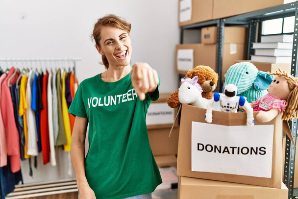 Hermosa mujer caucásica vistiendo una camiseta voluntaria en el stand de donaciones señalándote a ti y a la cámara con los dedos, sonriendo positiva y alegre  - Foto, imagen