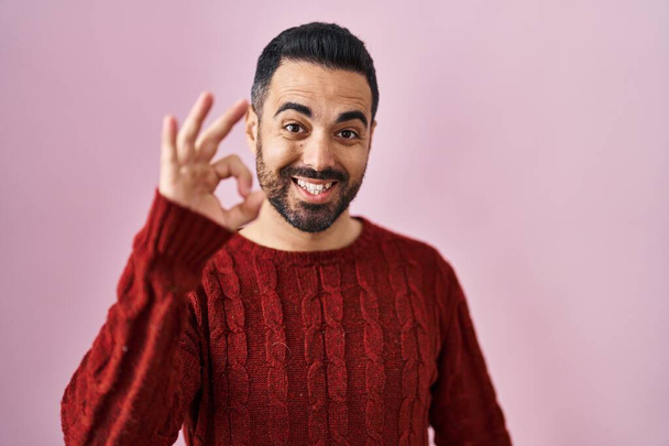 Νεαρός Ισπανός με γενειάδα φορώντας casual πουλόβερ πάνω από ροζ φόντο χαμογελώντας θετικά κάνει ok σημάδι με το χέρι και τα δάχτυλα. επιτυχής έκφραση.  - Φωτογραφία, εικόνα