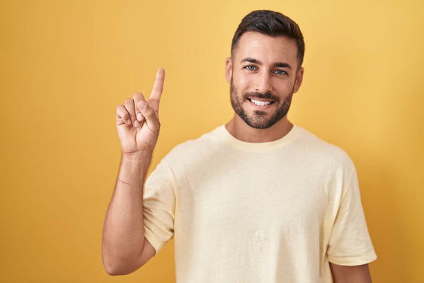 Όμορφος Ισπανόφωνος στέκεται πάνω από κίτρινο φόντο δείχνει και δείχνει προς τα πάνω με το δάχτυλο νούμερο ένα, ενώ χαμογελά αυτοπεποίθηση και χαρούμενος.  - Φωτογραφία, εικόνα
