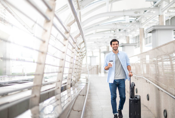 Человек, держащий чашку кофе и вытаскивающий багаж, пока он ждет своего друга в аэропорту. - Фото, изображение