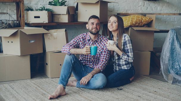 Girlfriend and boyfriend are talking making plans sitting on floor of new purchased apartment, looking around and holding mugs. Relationship, people and relocation concept. - Valokuva, kuva