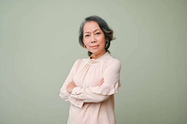 Уверенная и успешная женщина 60-х годов, отставная азиатка в повседневной одежде, скрестила руки, посмотрела в камеру и стояла изолированная на зеленом фоне студии. - Фото, изображение